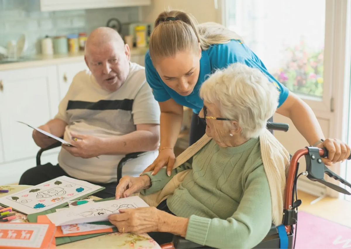 Woman assisting an elderly woman in a wheelchair with activities, with a man observing.