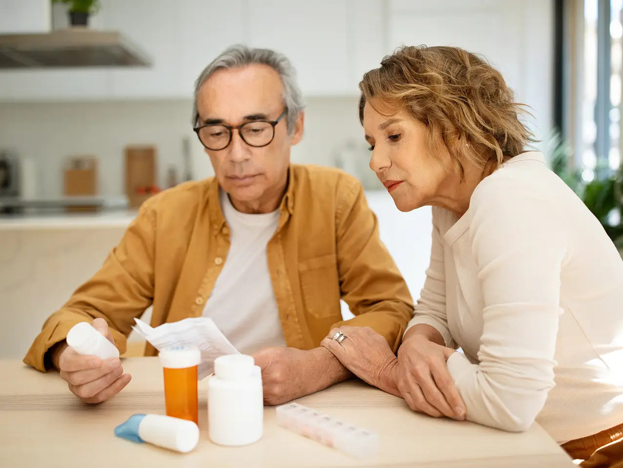 An older man and woman sitting at a table in a bright kitchen or medication-area setting, looking at a pill bottle and paper while several prescription pill bottles and a pill organizer are on the table.