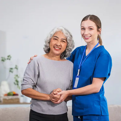 A smiling elderly woman with gray hair in a ribbed light gray top is being supported by a female caregiver in bright blue scrubs, standing in a brightly lit indoor setting.
