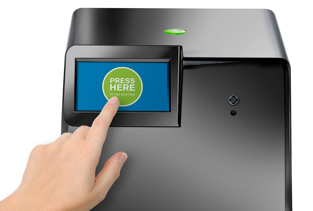 Shows a finger pressing the touch screen on the MedaCube. The screen shows a press here button showing that medications are ready to take. 